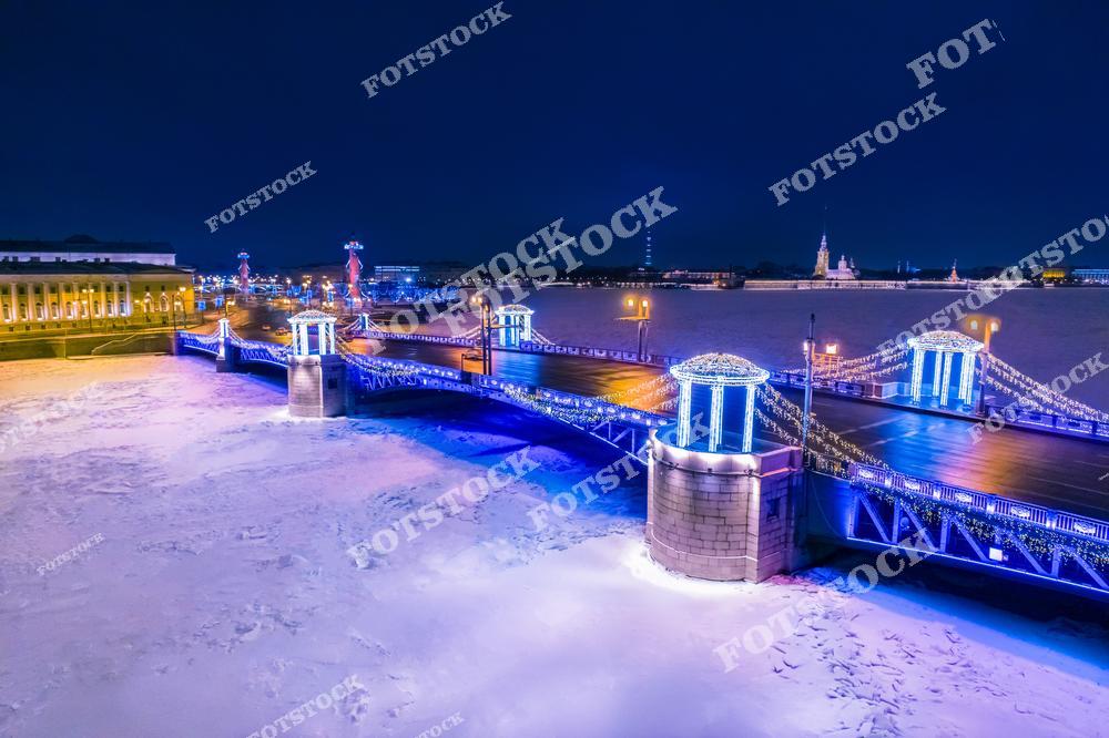На фотографии изображён Дворцовый мост, украшенный праздничной иллюминацией в зимнюю ночь. На левом берегу виднеются Ростральные колонны и здание Биржи на Стрелке Васильевского острова. На противоположной стороне — силуэт Петропавловской крепости с собором святых Петра и Павла. Нева скована льдом, подчёркивая морозную атмосферу. Светящиеся гирлянды и огни придают городу волшебный новогодний облик.
